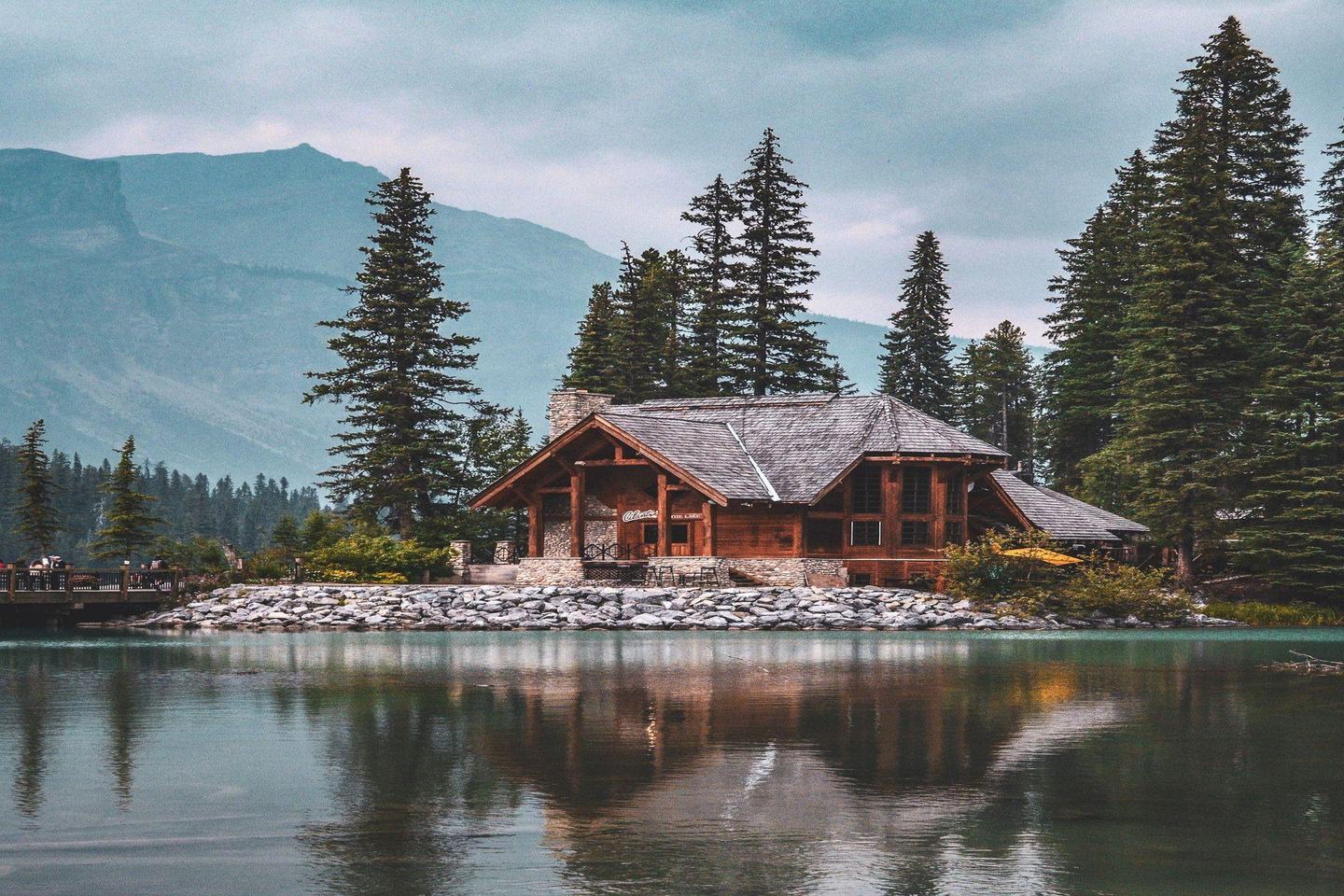 A log cabin on a lakeshore surrounded by conifers and mountains